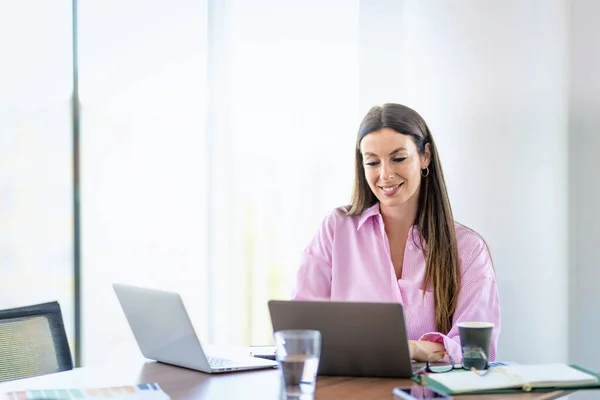 微笑的女商人坐在现代化的办公室里 用笔记本电脑工作 布鲁内特头发的职业女性微笑着 穿着衬衫 — 图库照片