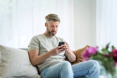 Orta yaşlı bir adam evdeki kanepede dinleniyor ve cep telefonu kullanıyor. Beyaz erkek, günlük kıyafetler giyiyor ve mesajlaşıyor.. 