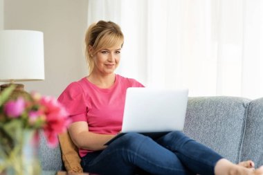 Sarı saçlı kadın koltukta oturuyor ve iş için dizüstü bilgisayar kullanıyor ya da video çekiyor. Kendine güvenen, sıradan kıyafetler giyen ve gülümseyen bir kadın. Merkez Ofis. 