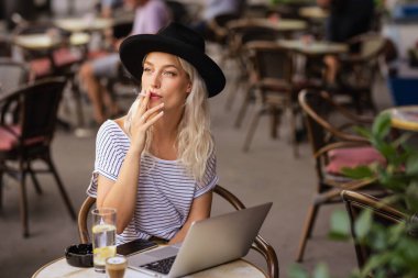 Sigara içen ve dizüstü bilgisayarını kullanan genç bir kadın tipik bir Fransız kafe terasında oturuyor. Sarı saçlı, şapkalı bir kadın. Sokak modasına uygun. 