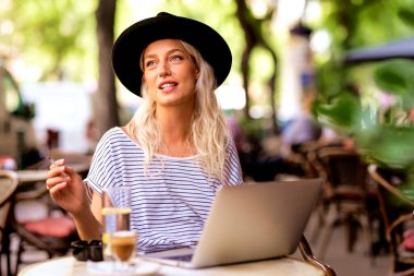 Sigara içen ve dizüstü bilgisayarını kullanan genç bir kadın tipik bir Fransız kafe terasında oturuyor. Sarı saçlı, şapkalı bir kadın. Sokak modasına uygun. 