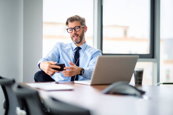 Portrait of businessman using smartphone and earphones while sitting at the office and having a video call. Professional man wearing shirt and tie. 