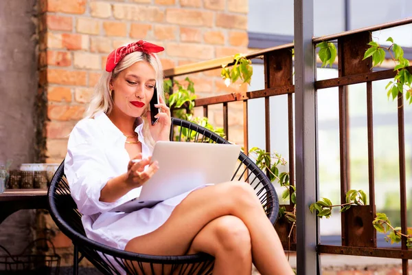 自宅のバルコニーでラップトップで作業している間 魅力的な女性の喫煙タバコと電話を持っています ブロンドの髪の女性はパリッシュスタイルの頭のスカーフと特大の白いシャツを着て — ストック写真