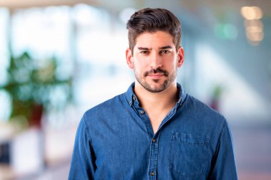 Kendine güveni tam, orta yaşlı, sıradan kıyafetler giyen ve içeride duran bir adam. Mavi gömlek giyiyor ve kameraya bakıyor..