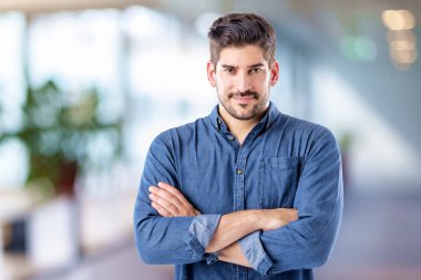 Kendine güveni tam, orta yaşlı, sıradan kıyafetler giyen ve içeride duran bir adam. Mavi gömlek giyiyor ve kameraya bakıyor..