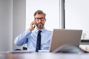 Ofis masasında oturan ciddi yüzlü bir iş adamı iş için dizüstü bilgisayar ve cep telefonu kullanıyor. Orta yaşlı erkek profesyonel iş görüşmesi yapıyor..