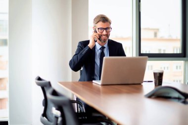 Yetişkin bir iş adamı, dizüstü bilgisayarla masada oturuyor ve cep telefonuyla konuşuyor. Ofiste çalışan bir iş adamı. Takım elbiseli ve kravatlı profesyonel bir adam.. 