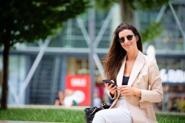 Çekici esmer saçlı kadın sokakta bir bankta oturuyor ve telefon kullanıyor. İş kıyafeti, güneş gözlüğü ve kısa mesaj takan gülümseyen bir kadın.. 