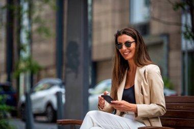 Çekici kahverengi saçlı kadın sokakta bir bankta oturuyor ve telefon kullanıyor. Neşeli gülümseyen kadın iş kıyafetleri ve mesajlar takıyor.. 