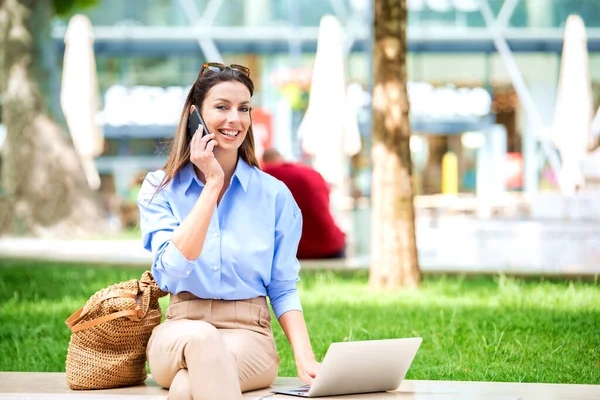 魅力的な茶色の髪の女性は 携帯電話で話して ノートパソコンを使用して通りのベンチに座っている 陽気な笑顔の女性はカジュアルな服を着てインターネット上で働く — ストック写真