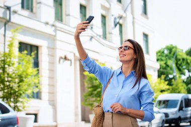 Esmer kadın caddede yürüyor ve akıllı telefonuyla selfie çekiyor. Gülen kadın güneş gözlüğü ve günlük kıyafetler takıyor..