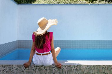 Yüzme havuzunda dinlenirken hasır şapka ve pembe gömlek giyen bir kadın..