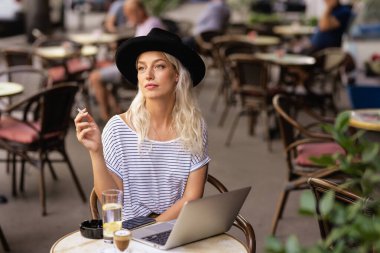 Sigara içen ve dizüstü bilgisayarını kullanan genç bir kadın tipik bir Fransız kafe terasında oturuyor. Sarı saçlı, şapkalı bir kadın. Sokak modasına uygun. 