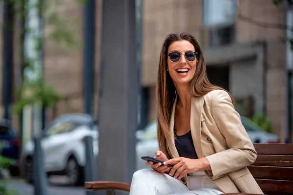 笑着棕色头发的女人坐在街上的长椅上 用电话 穿着商务休闲装和发短信的迷人女性 — 图库照片