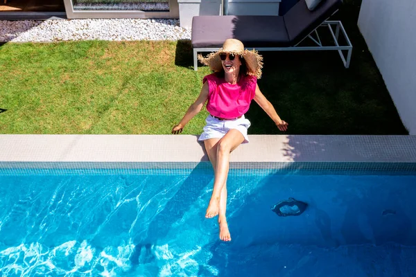 stock image Full length shot of a cheerful woman wearing straw hat and pink t-shirt while relaxing at swimming pool.