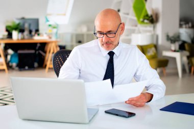 Finans işadamı masasında oturuyor, dizüstü bilgisayar kullanıyor ve elinde belgeler tutuyor. Finansal bir açıklama yapıyor. Gömlek ve kravat giyen profesyonel bir adam..  
