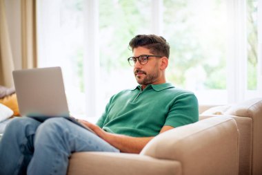 Orta yaşlı bir adam evdeki koltuğunda oturuyor ve iş için dizüstü bilgisayar kullanıyor. Merkez Ofis.