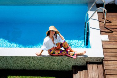 Happy woman wearing white shirt and straw hat while relaxing by the poolside. Full length shot. Copy space. 