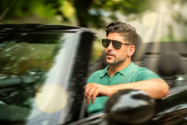 Güneş gözlüğü takan, üstü açık bir arabada oturan ve yolda araba süren yakışıklı bir adam.. 