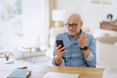 Evde masada oturan ve elinde akıllı telefon tutan şaşırmış bir adam. Kendine güveni tam bir erkek, merak uyandıran bir yüz mesajı ya da video çağrısı var. Merkez Ofis.