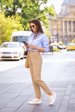 Uzun boylu, kahverengi saçlı, sıradan elbiseli bir kadın sokakta duruyor ve mesajlaşıyor. Güneş gözlüğü takan ve gülümseyen çekici bir kadın.. 