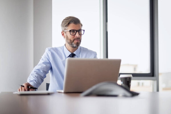 Бизнесмен средних лет сидит за своим столом в офисе, используя ноутбук для работы. Исполнительный профессионал в очках и рубашке. 