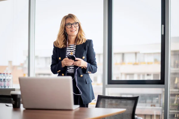 Mid aged businesswoman standing in a modern office and having video call. Confident professional woman using earphone and smartphone and having video call.