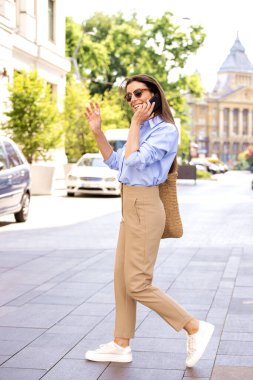 Çekici bir kadın sokakta yürürken taksi çağırmak ve telefon etmek için elini kaldırıyor. Gömlek ve güneş gözlüğü takan esmer bir kadın. Tam boy çekim. 