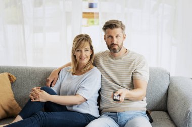 Mutlu bir orta yaşlı çiftin evde televizyon izlemesi. Sarı saçlı kadın ve sıradan kıyafetler giyen yakışıklı adam kanepede oturuyor. Uzaktan kumandayı elinde tutan adam. 