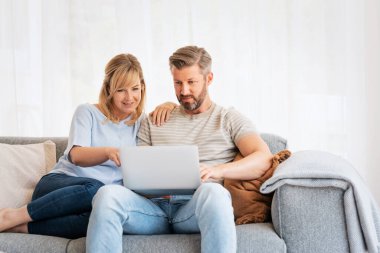 Orta yaşlı bir çift kanepede oturuyor ve dizüstü bilgisayar kullanıyorlar. Sarı saçlı kadın ve gri saçlı adam internette sörf yapıyor ya da video çekiyor.. 