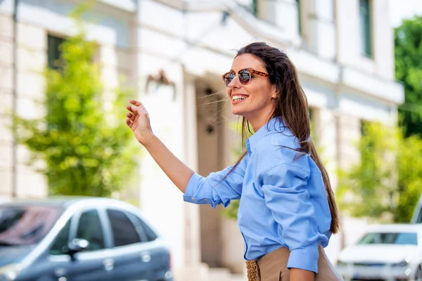 一个迷人的女人在街上走的时候举手叫出租车 布鲁内特带着蓝色衬衫和太阳镜的女性头发 — 图库照片