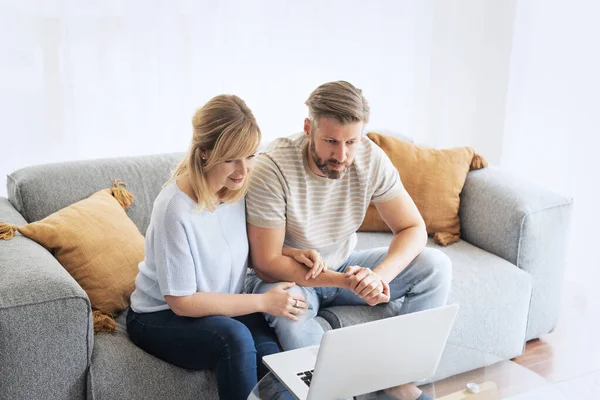 Middle-aged couple sitting at home on the sofa and using a laptop. Blond haired woman and grey haired man surfing the internet or having video call.