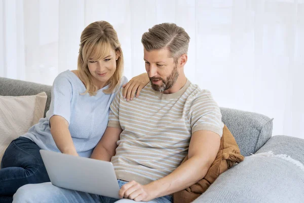 Middle-aged couple sitting at home on the sofa and using a laptop. Blond haired woman and grey haired man surfing the internet or having video call.