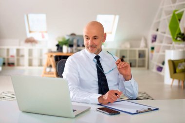 Orta yaşlı bir işadamı ofisinde oturuyor ve dizüstü bilgisayarı üzerinde çalışıyor. Profesyonel erkek web konferansı yapıyor.. 