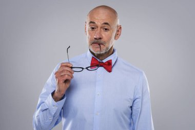 Gri arka planda duran orta yaşlı bir adam düşünceli görünüyor. Tişört ve papyon takan ve elinde cam tutan kendinden emin bir erkek. Boşluğu kopyala. 