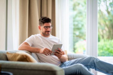 Orta yaşlı bir adam elinde dijital tabletle kanepede oturuyor. Gözlüklü ve günlük kıyafetler giyen kendinden emin bir erkek. Görüntülü arama yapıyor ya da internette geziniyor. Merkez Ofis.