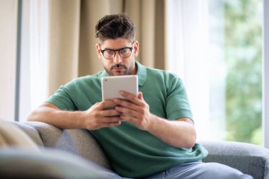 Orta yaşlı bir adam elinde dijital tabletle kanepede oturuyor. Gözlüklü ve günlük kıyafetler giyen kendinden emin bir erkek. Görüntülü arama yapıyor ya da internette geziniyor. Merkez Ofis.