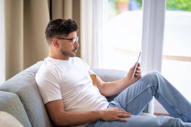 Orta yaşlı bir adam elinde dijital tabletle kanepede oturuyor. Gözlüklü ve günlük kıyafetler giyen kendinden emin bir erkek. Görüntülü arama yapıyor ya da internette geziniyor. Merkez Ofis.