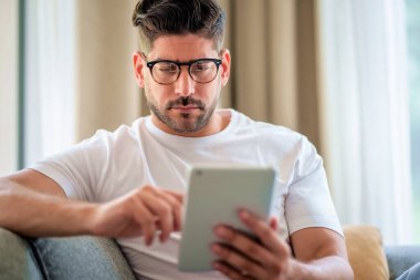 Orta yaşlı bir adamın kanepede oturup elinde dijital tablet tutmasının yakın çekimi. Gözlüklü ve günlük kıyafetler giyen kendinden emin bir erkek. Görüntülü arama yapıyor ya da internette geziniyor. Merkez Ofis.
