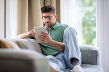 Orta yaşlı bir adam elinde dijital tabletle kanepede oturuyor. Gözlüklü ve günlük kıyafetler giyen kendinden emin bir erkek. Görüntülü arama yapıyor ya da internette geziniyor. Tam boy çekim. 