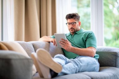 Orta yaşlı bir adam elinde dijital tabletle kanepede oturuyor. Gözlüklü ve günlük kıyafetler giyen kendinden emin bir erkek. Görüntülü arama yapıyor ya da internette geziniyor. Tam boy çekim. 