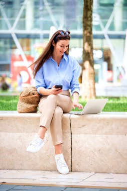 Şehirdeki bir bankta oturan esmer bir kadın iş için dizüstü bilgisayar ve akıllı telefon kullanıyor. Gündelik kıyafet olarak güneş gözlüğü takan orta yaşlı mutlu bir kadın. Tam boy çekim. 