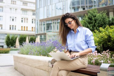 Şehirdeki bir bankta oturan esmer saçlı kadın ve iş için dizüstü bilgisayar kullanıyor. Mutlu orta yaşlı kadın güneş gözlüğü ve günlük kıyafetler takıyor.. 