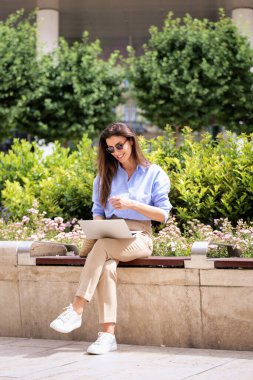 Şehirdeki bir bankta oturan esmer saçlı kadın ve iş için dizüstü bilgisayar kullanıyor. Mutlu orta yaşlı kadın güneş gözlüğü ve günlük kıyafetler takıyor. Tam boy çekim. 
