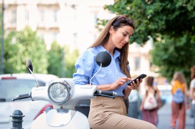 Scooter 'ı şehir caddesinde duran ve cep telefonundan mesaj atan gülen bir kadın. Esmer, kadın, mavi gömlek giyiyor.. 