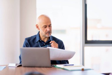 Orta yaşlı iş adamı modern bir ofiste masada oturuyor ve dizüstü bilgisayar kullanıyor. Profesyonel erkek düşünceli görünüyor ve evrak işleriyle uğraşıyor.. 