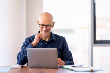 Orta yaşlı bir işadamı modern bir ofiste masa başında oturuyor ve dizüstü bilgisayarını iş için kullanıyor. Profesyonel erkek video görüşmesi yapıyor.. 