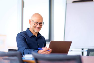 Orta yaşlı bir işadamı modern bir ofiste masa başında oturuyor ve dizüstü bilgisayarını iş için kullanıyor. Profesyonel erkek video görüşmesi yapıyor.. 