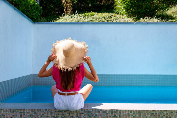 A woman sitting with her back to the pool. Relaxed female wearing pink shirt and straw hat. Full length shot. Copy space. 