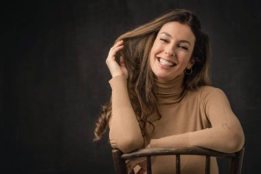 Çekici, orta yaşlı, dişlek, balıkçı yaka kazak giyen ve izole, koyu arka planda oturan bir kadının yakın çekimi. Uzayı kopyala Stüdyo çekimi. Elini alnına koy..